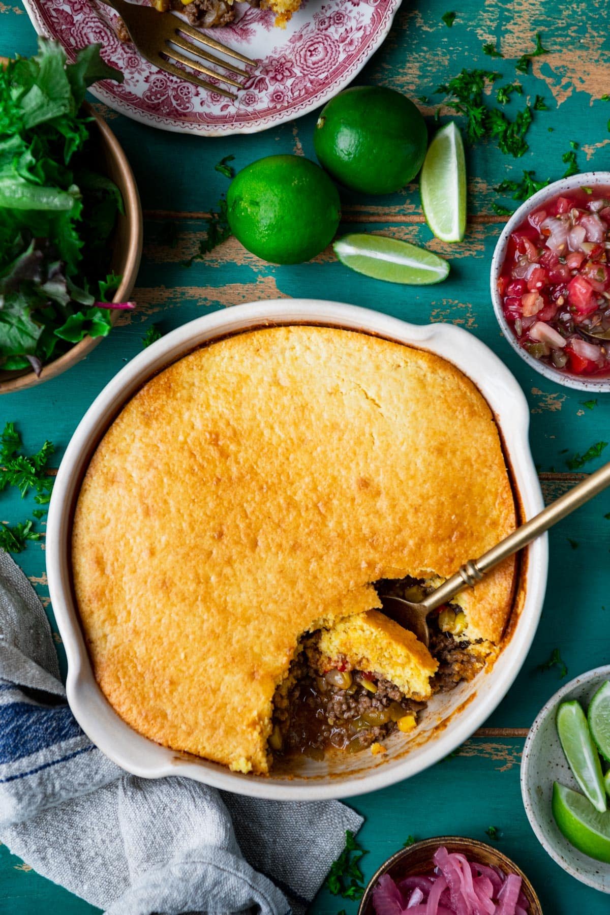 Overhead shot of a taco pie recipe surrounded by salad, fresh limes, and salsa.