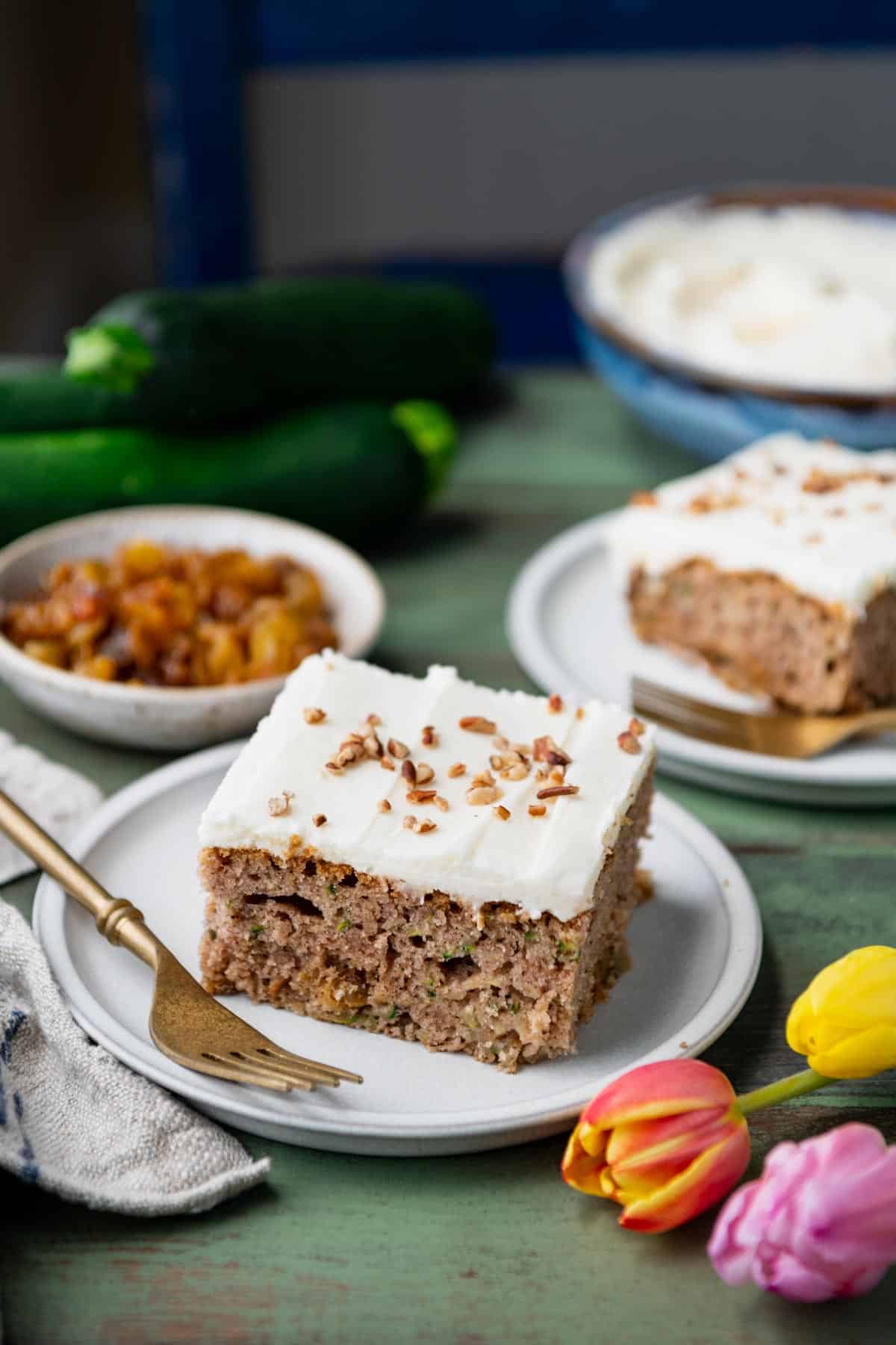 Two slices of zucchini cake on a table with raisins and cream cheese frosting in the background.