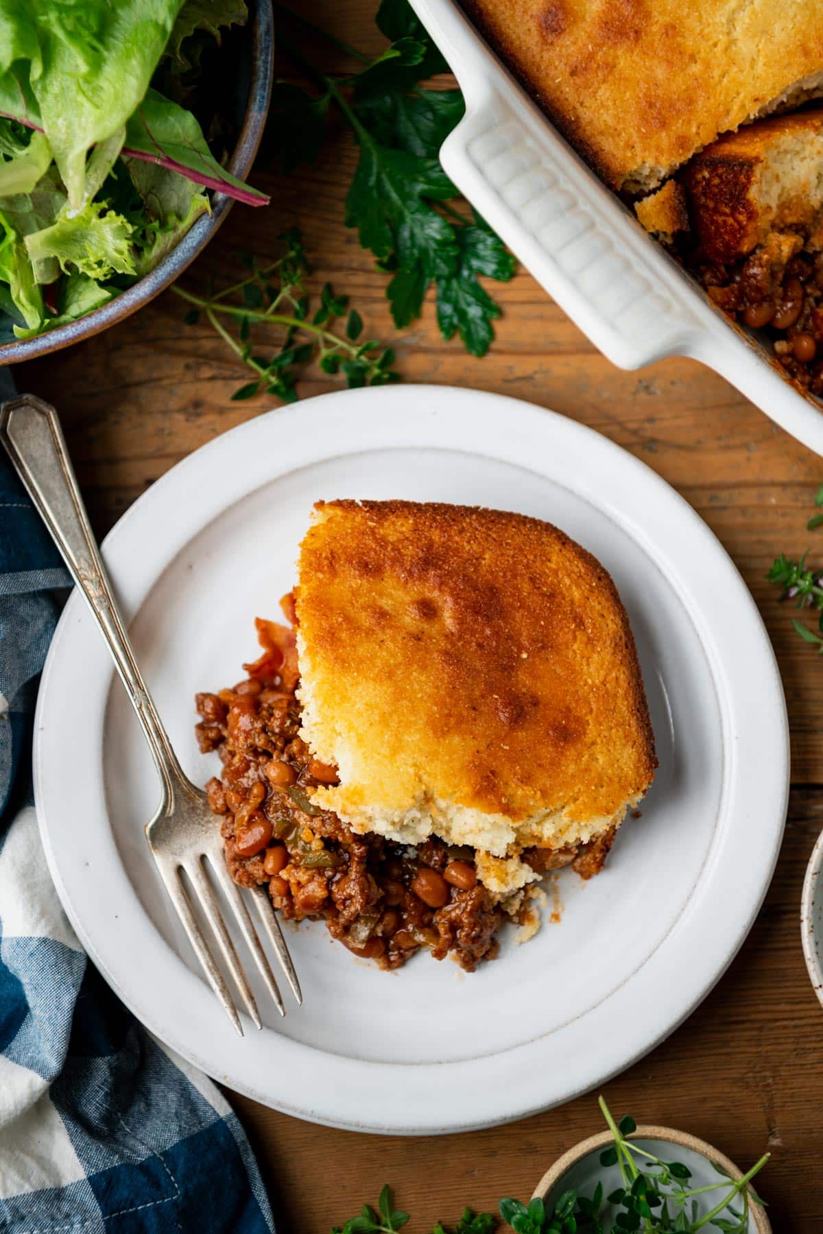 Overhead image of a bbq ground beef cornbread casserole on a wooden table with a side salad.