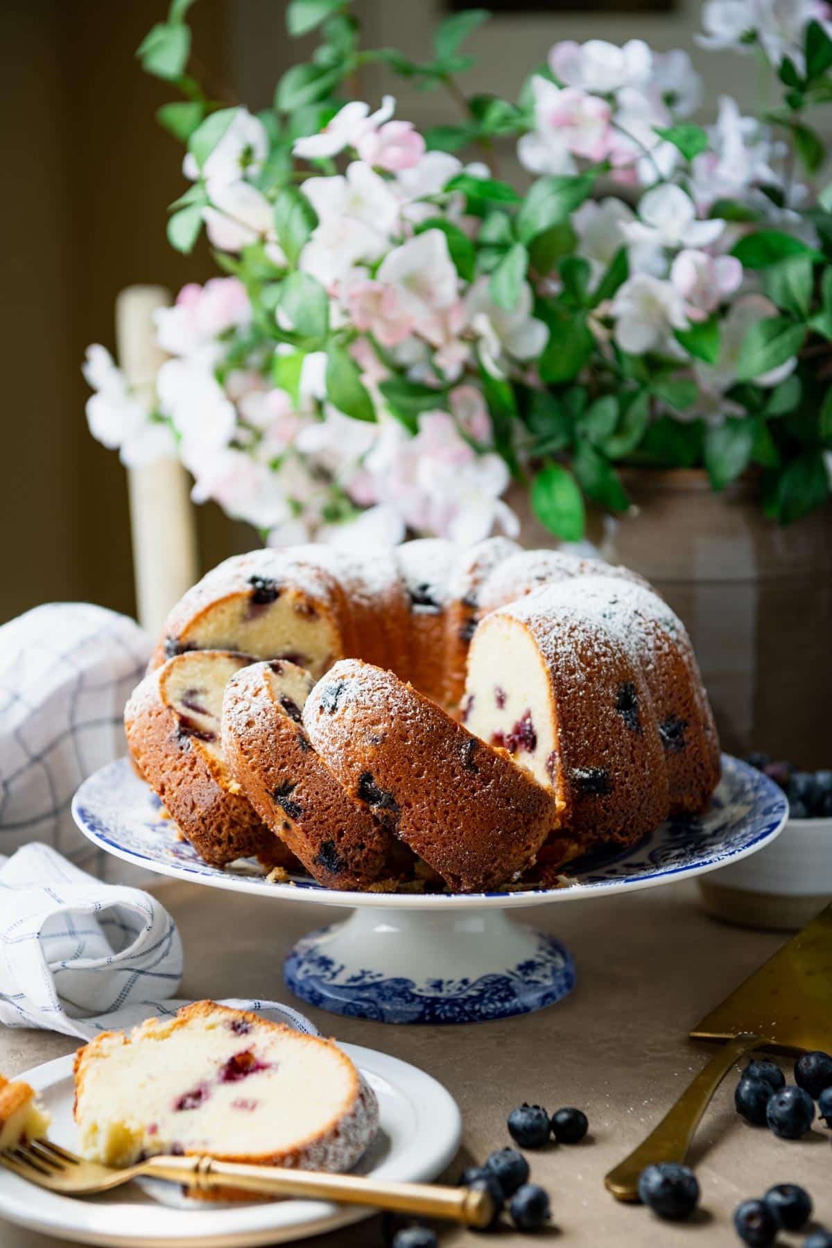 Sliced cream cheese pound cake with blueberries on a table with flowers in the background.