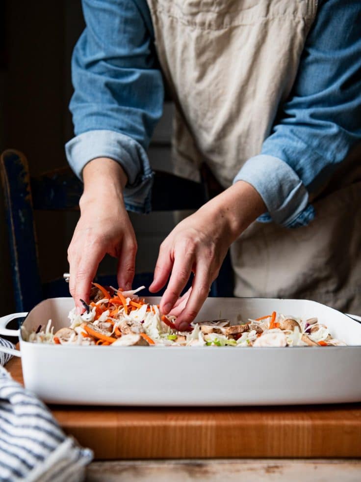 Stirring together coleslaw mix, carrots, mushrooms, and other veggies in a casserole dish.