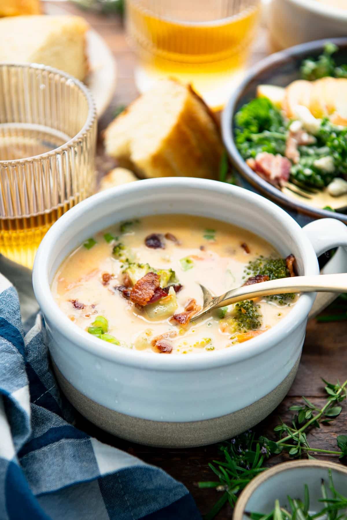 Side shot of a gold spoon in a white bowl of beer cheese soup with bacon and broccoli.
