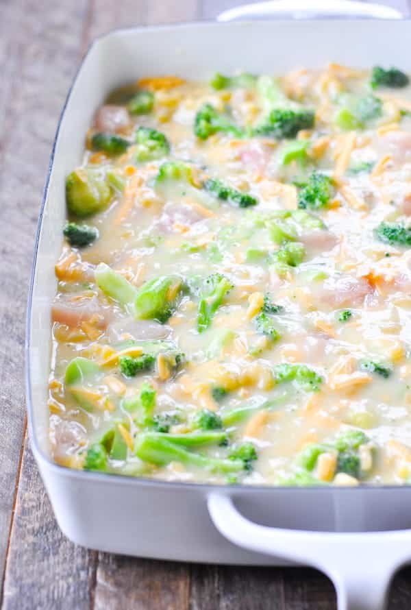 All of the uncooked ingredients for cheesy chicken broccoli pasta casserole -- a creamy soup mix, cubed chicken, broccoli, and cheese - poured into a white and blue casserole dish.