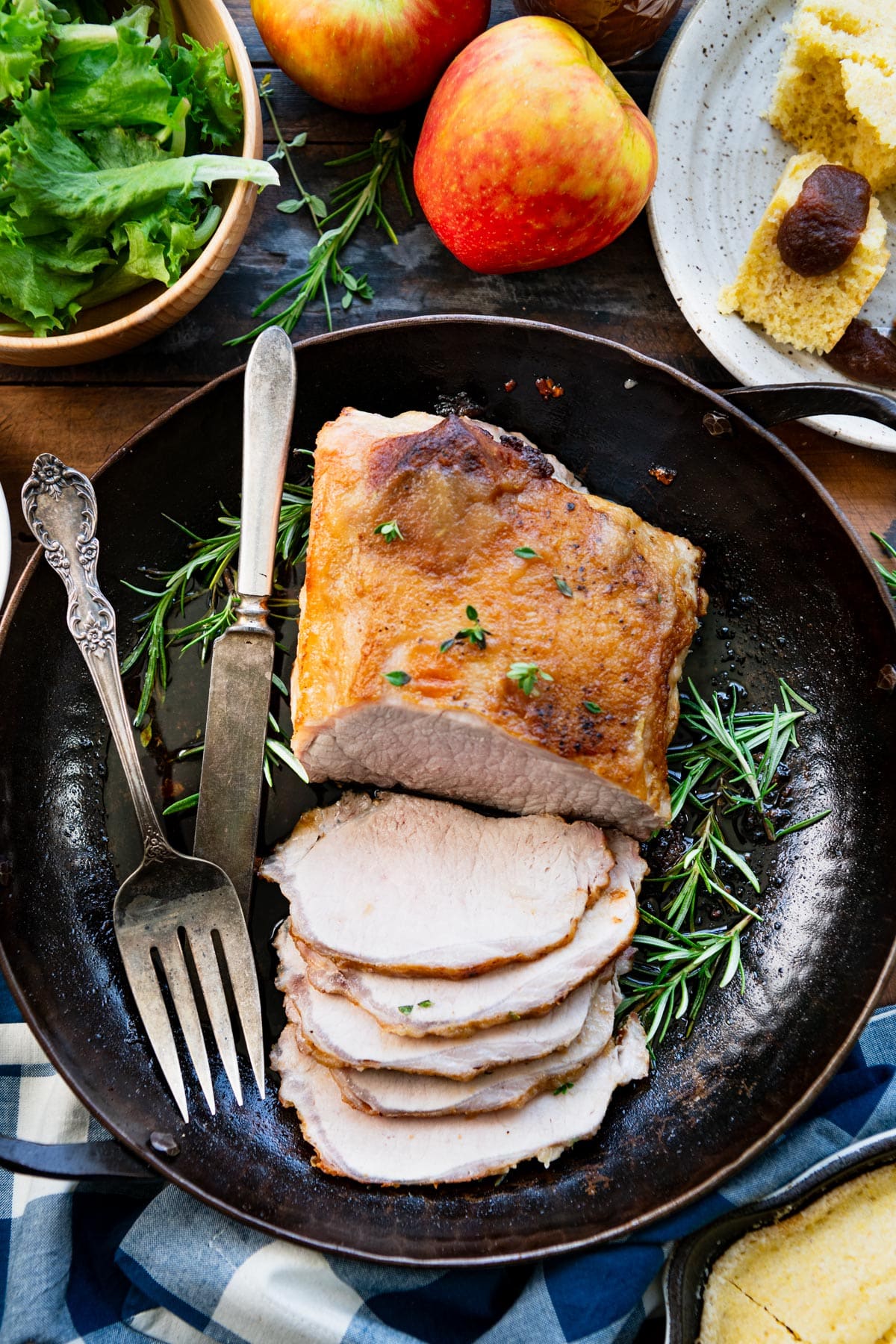 Overhead shot of a sliced pork loin on a wooden table with salad and cornbread.