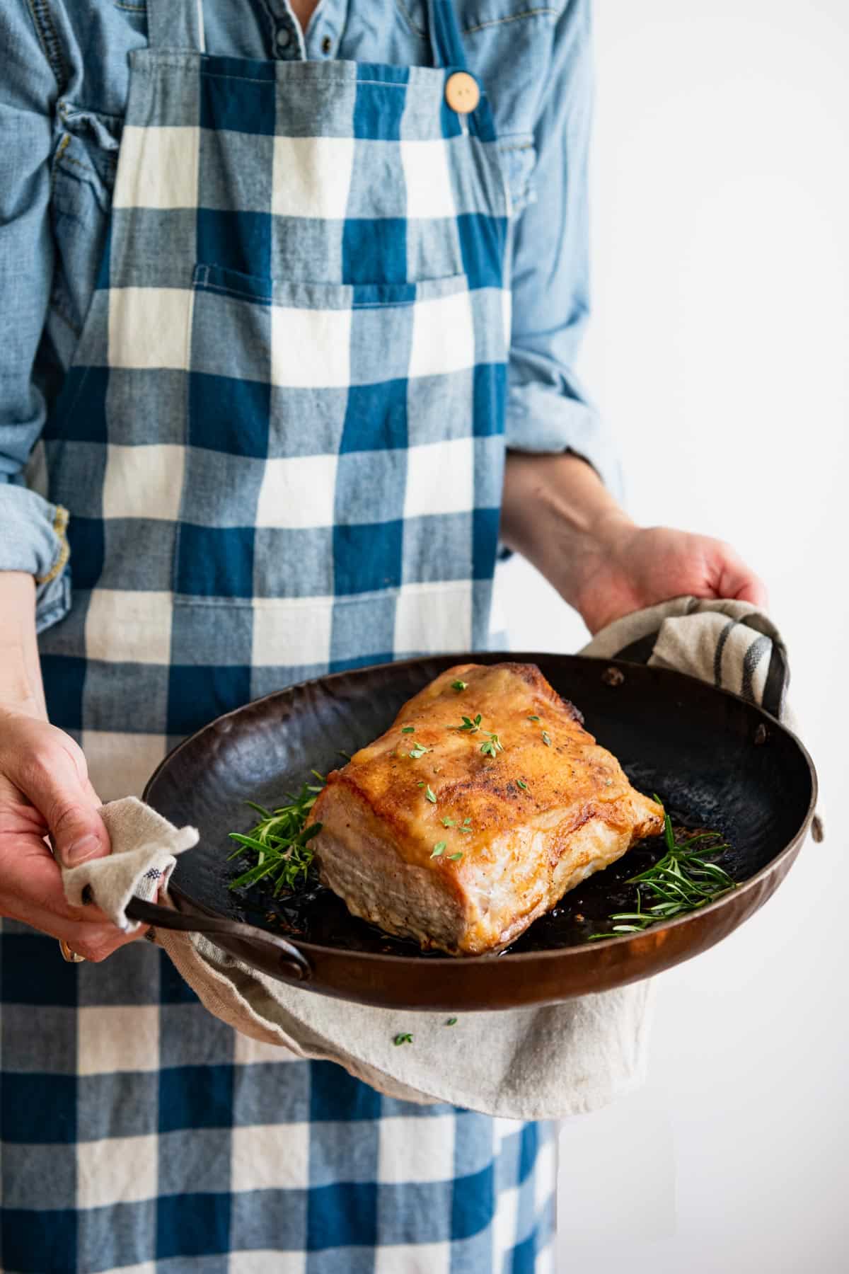 Cook in a blue and white apron holding a cast iron pan with an oven roasted pork loin.