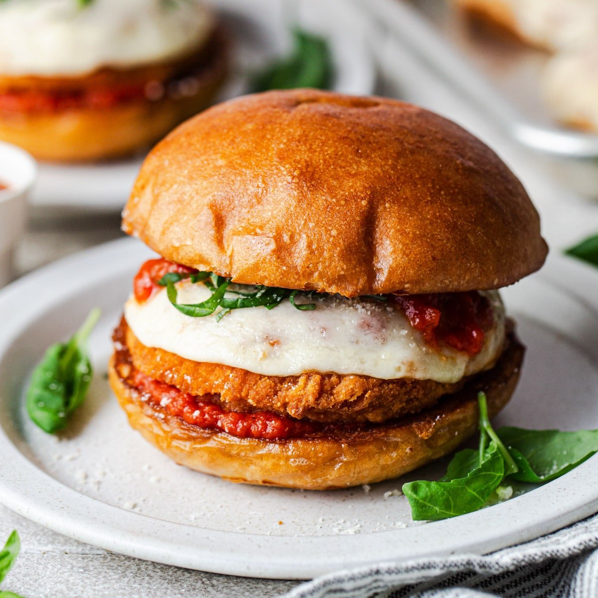 Square side shot of an easy chicken Parmesan sandwich on a white plate with fresh basil for garnish.