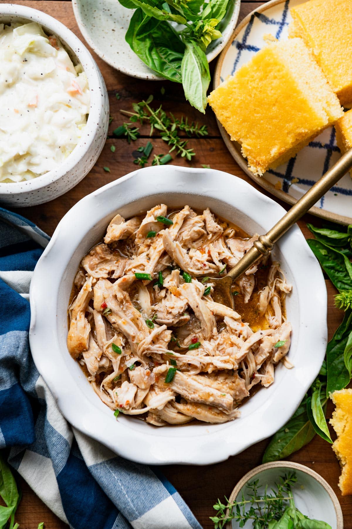 Slow cooker pulled chicken in a white bowl on a table with a side of cornbread and coleslaw.