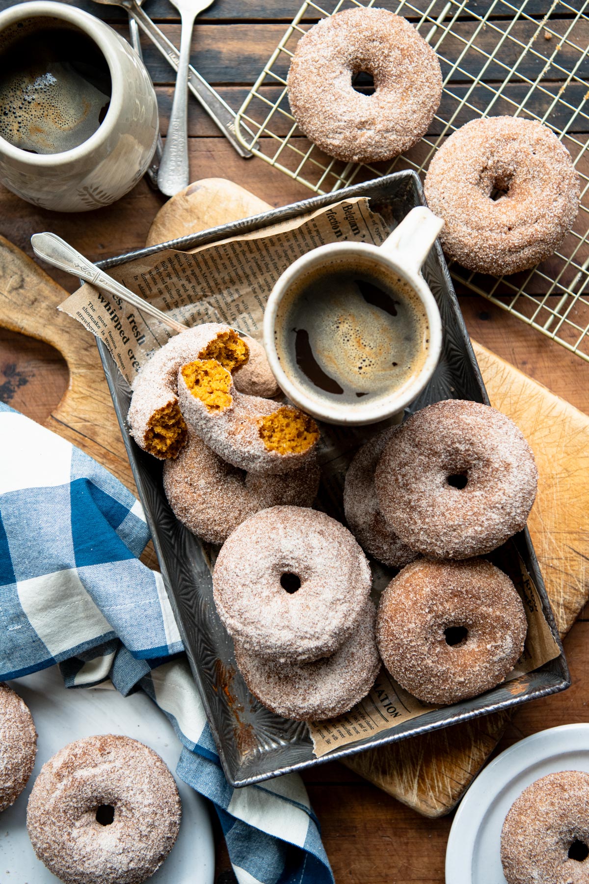 Wooden table full of a tray of pumpkin doughnuts with a blue and white check napkin nearby.