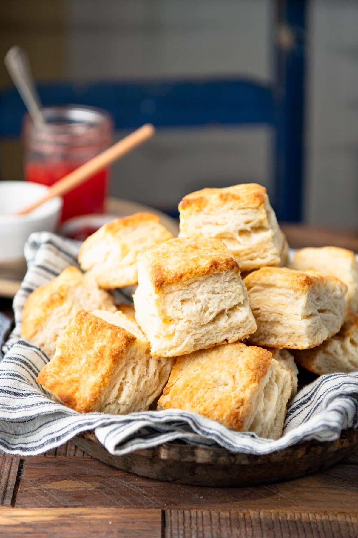Side shot of a basket of flaky biscuits on a wooden table with jam and honey in the background.
