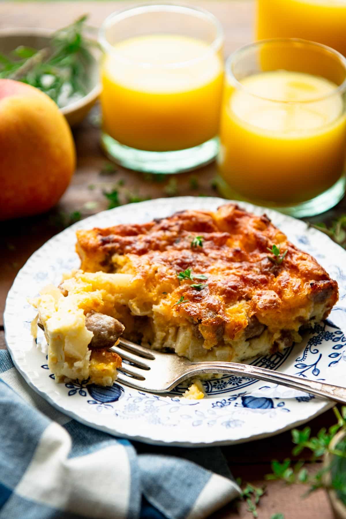 Side shot of a slice of apple and egg casserole on a plate with orange juice in the background.