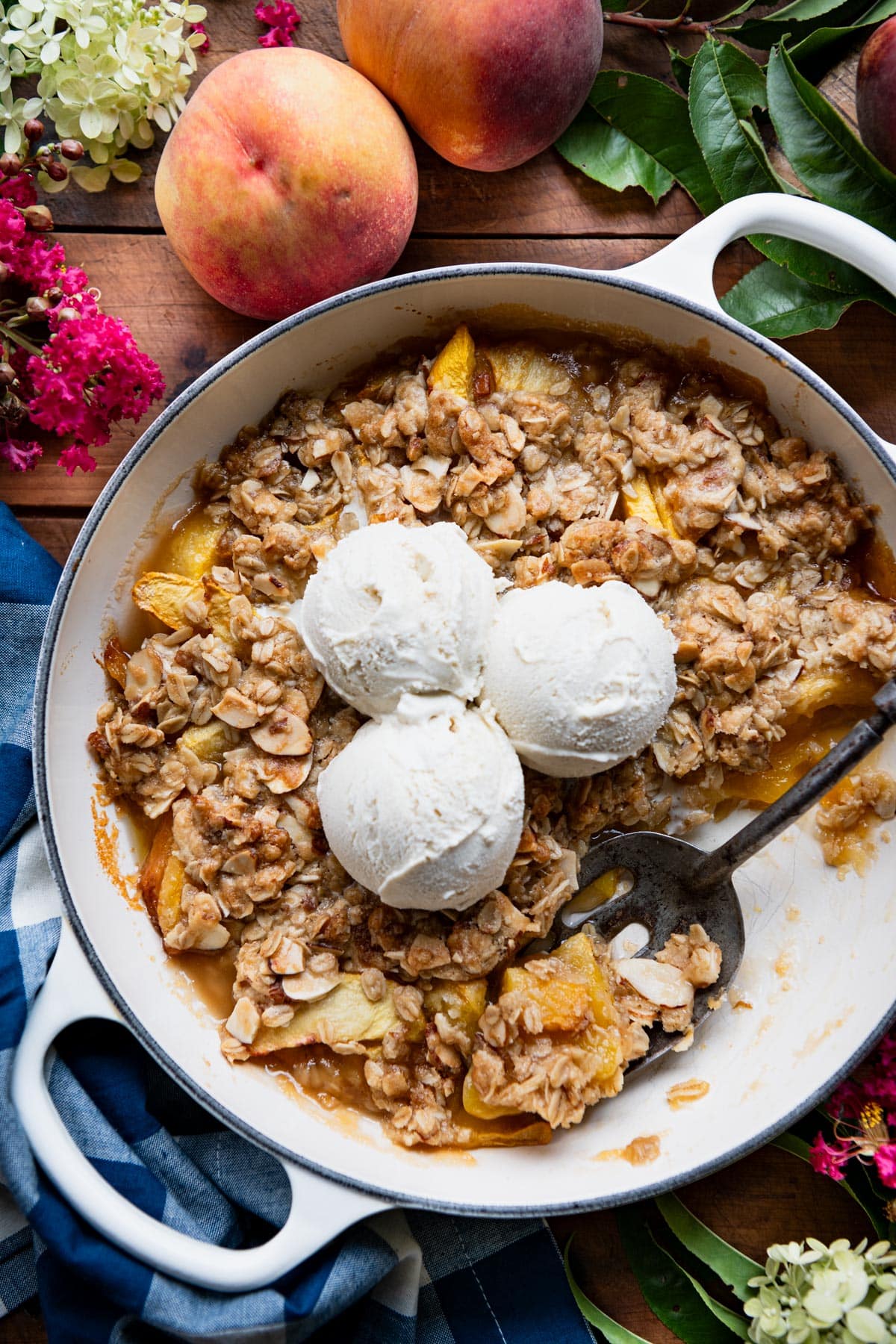 Overhead shot of an easy peach crisp recipe on a wooden table with ice cream and fresh flowers.