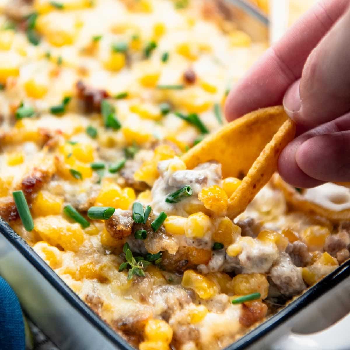 Square close up shot of hand using a chip to scoop up sausage dip.