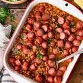 Pan of frank and beans on a wooden table with a serving spoon.