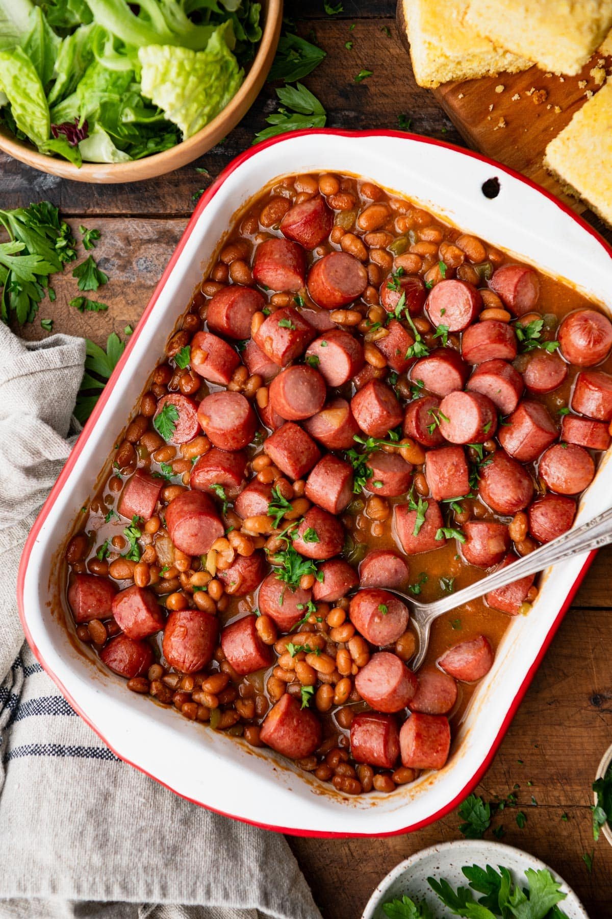 Pan of frank and beans on a wooden table with a serving spoon.