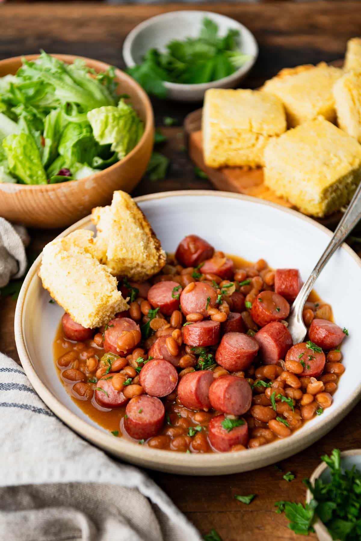 Side shot of a bowl of beans and franks on a table with a salad and cornbread in the background.