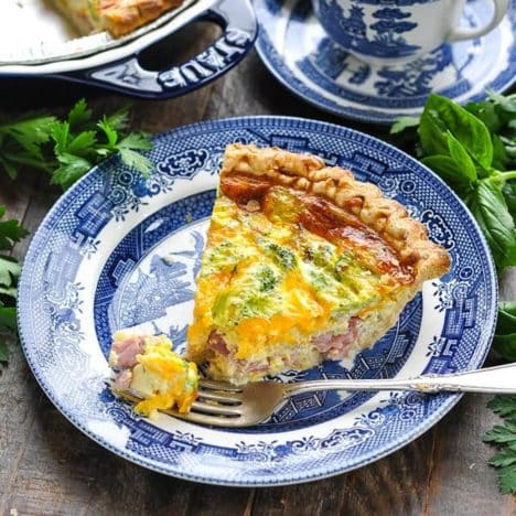 Square image of a slice of ham and broccoli quiche on a blue and white plate.