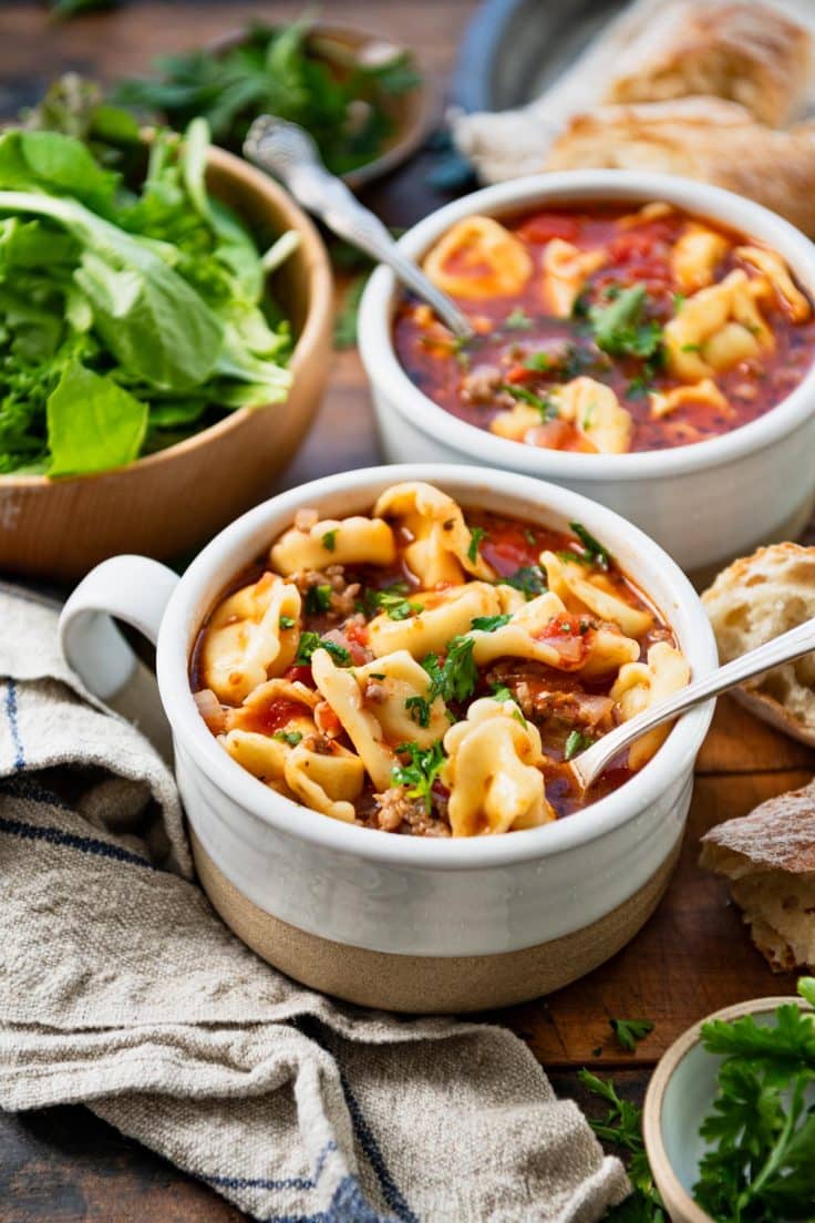 Close up front shot of two bowls of sausage tortellini soup on a wooden table with salad and bread.