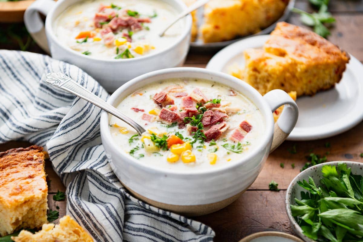 Horizontal shot of a bowl of crock pot chicken corn soup on a dinner table with cornbread.