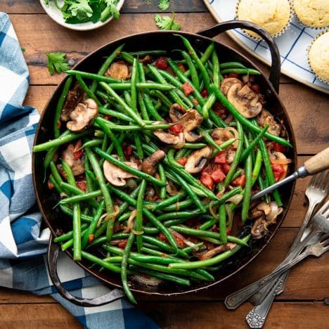 Square overhead shot of green beans and mushrooms in a pan on a wooden table with a side of cornbread muffins.