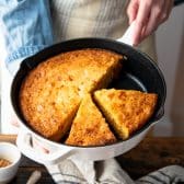 Square side shot of hands holding moist cornbread in a cast iron skillet.