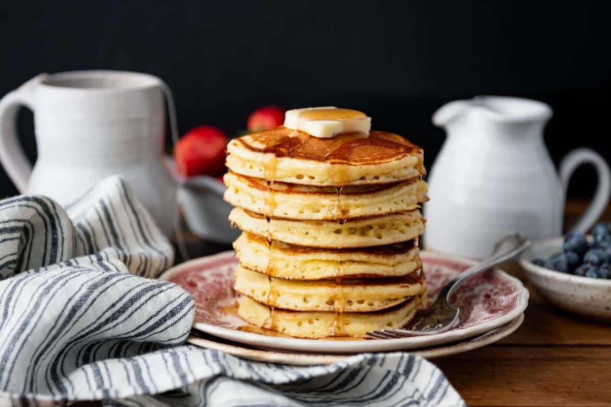Horizontal side shot of jiffy cornmeal pancakes on a breakfast table with syrup and tea in the background.