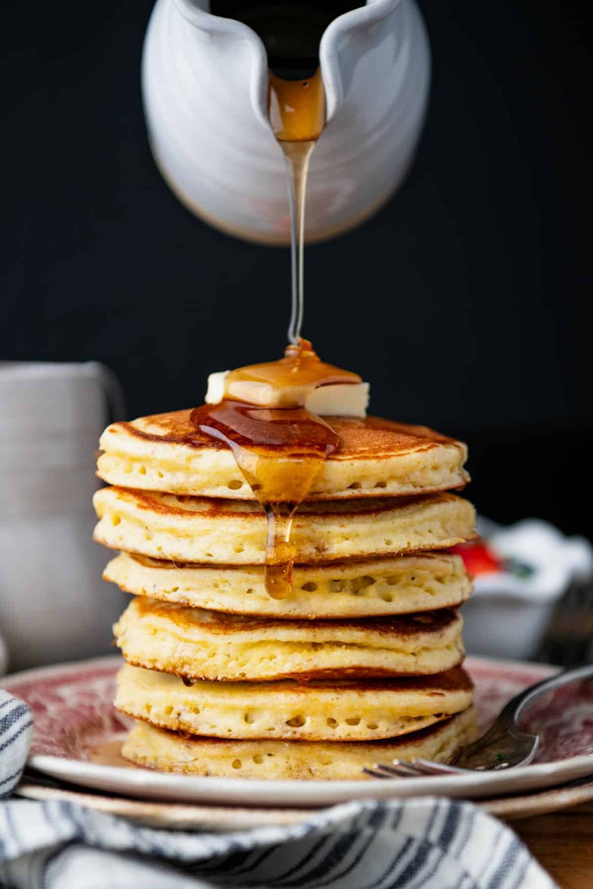 Pouring syrup over a plate of Jiffy cornmeal pancakes.