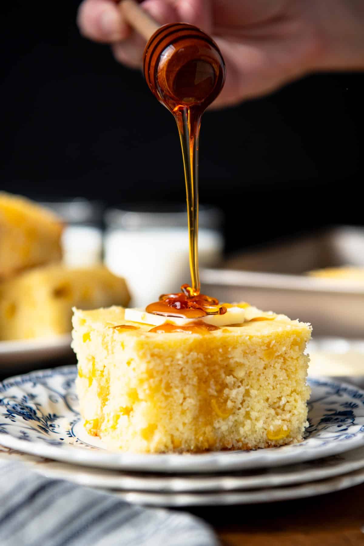 Drizzling honey over a slice of cornbread with corn on a blue and white plate.