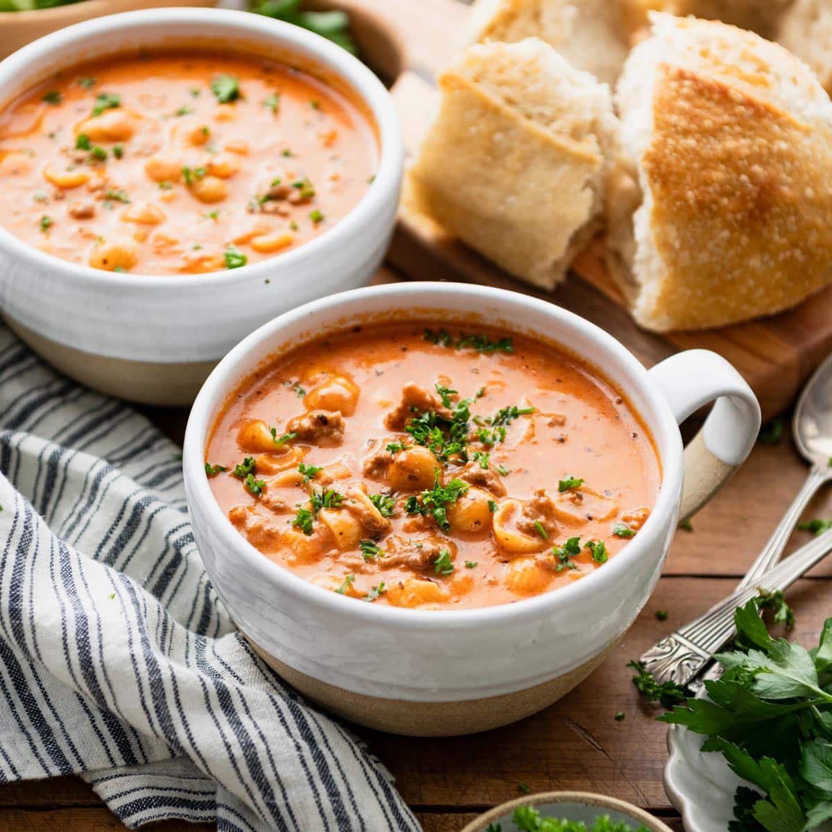 Square side shot of two bowls of creamy tomato soup with ground beef and noodles on a table with bread and salad.