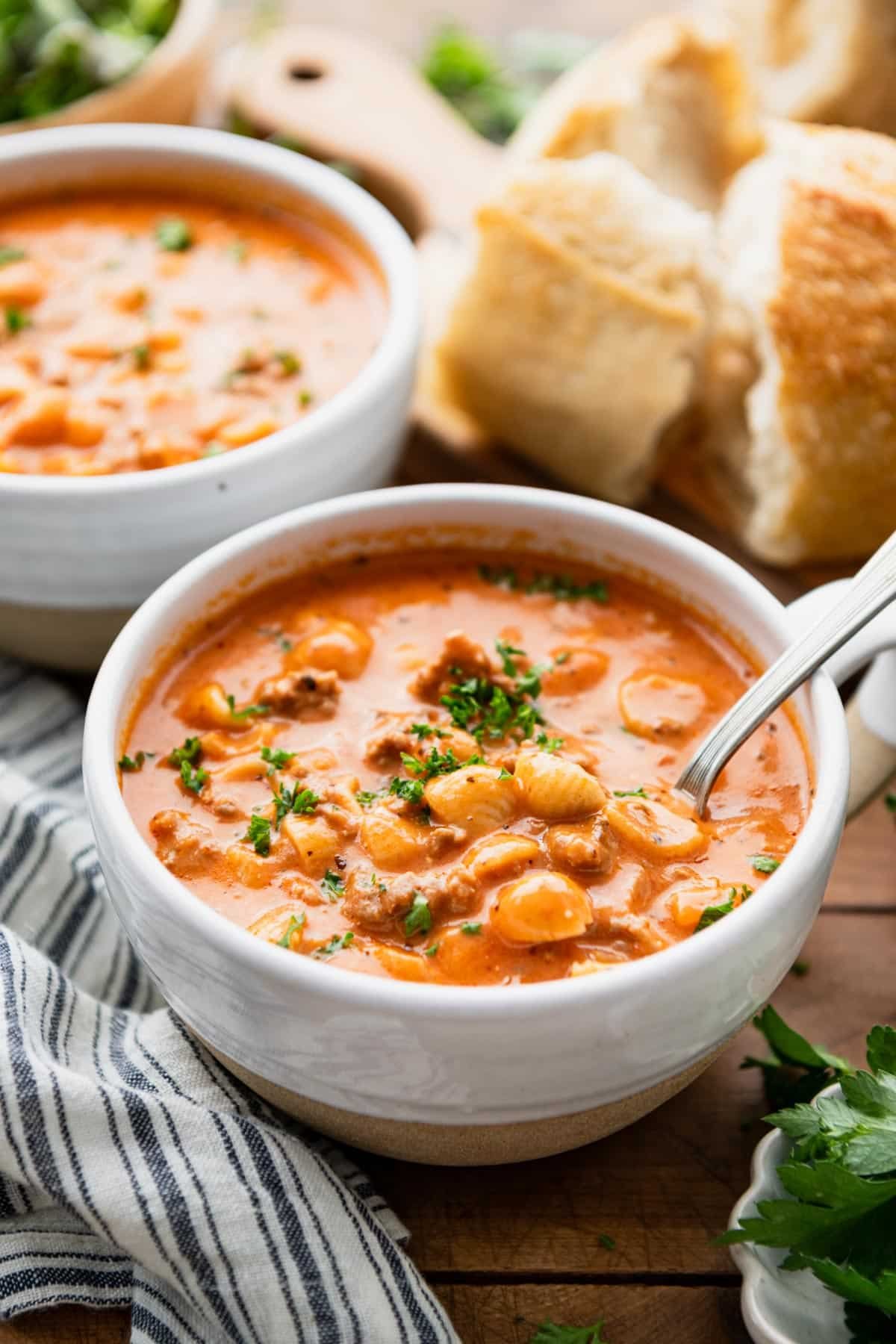 Side shot of two bowls of creamy beef tomato soup with noodles on a table with bread in the background.
