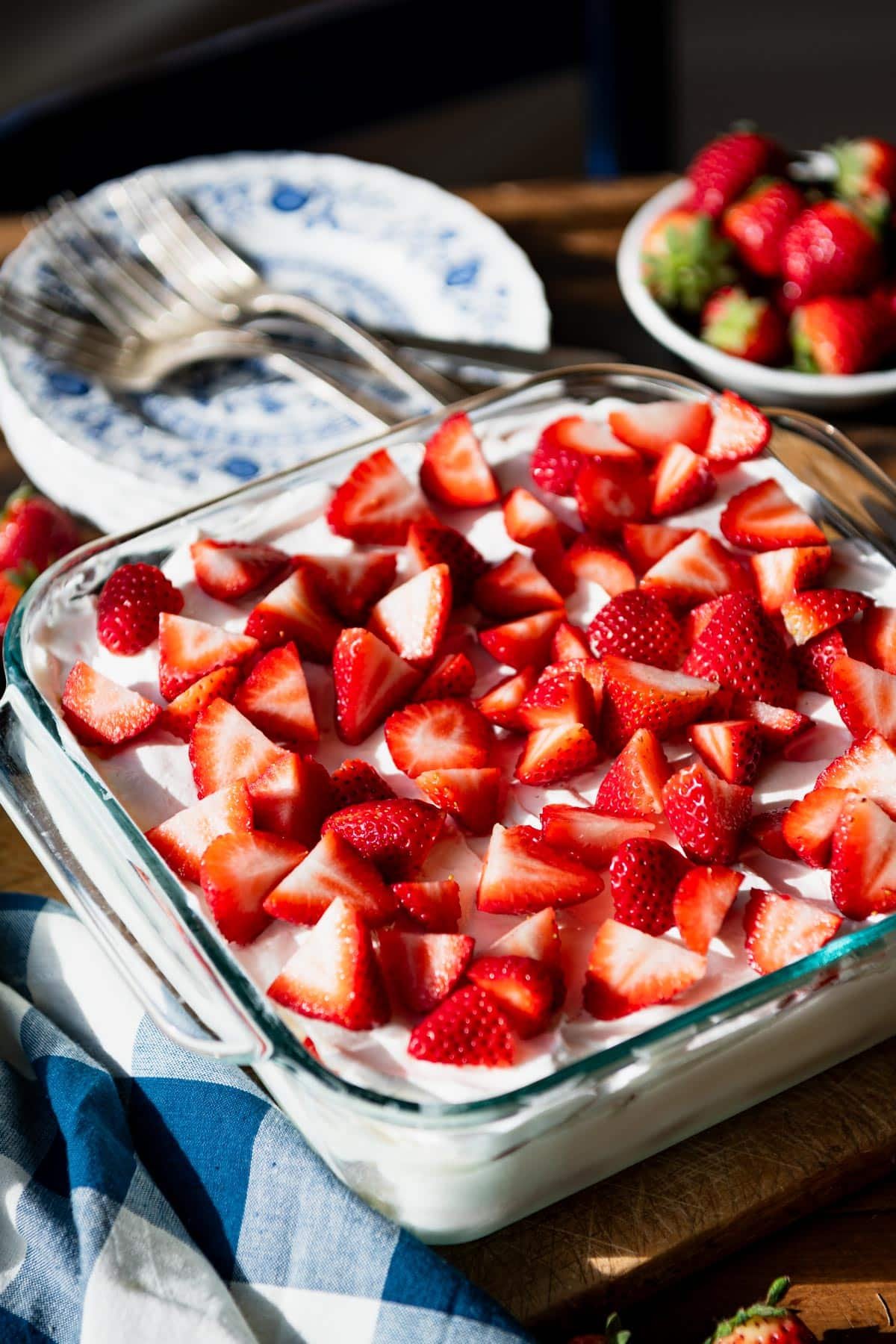 Close up side shot of a glass dish full of the best strawberry icebox cake recipe on a wooden table with a bowl of fresh berries in the background.