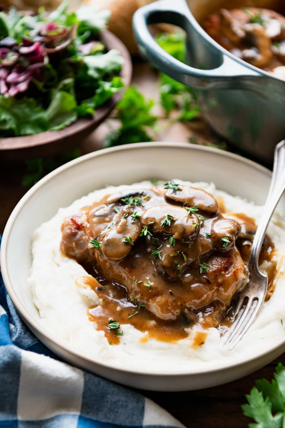 Side shot of a bowl of mashed potatoes topped with dutch oven pork chops and mushroom gravy.