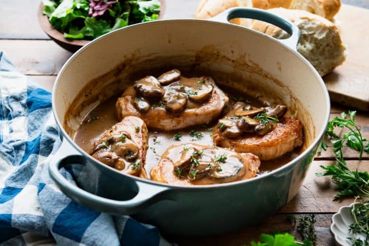 Horizontal side shot of a blue dutch oven with pork chops and mushroom gravy.