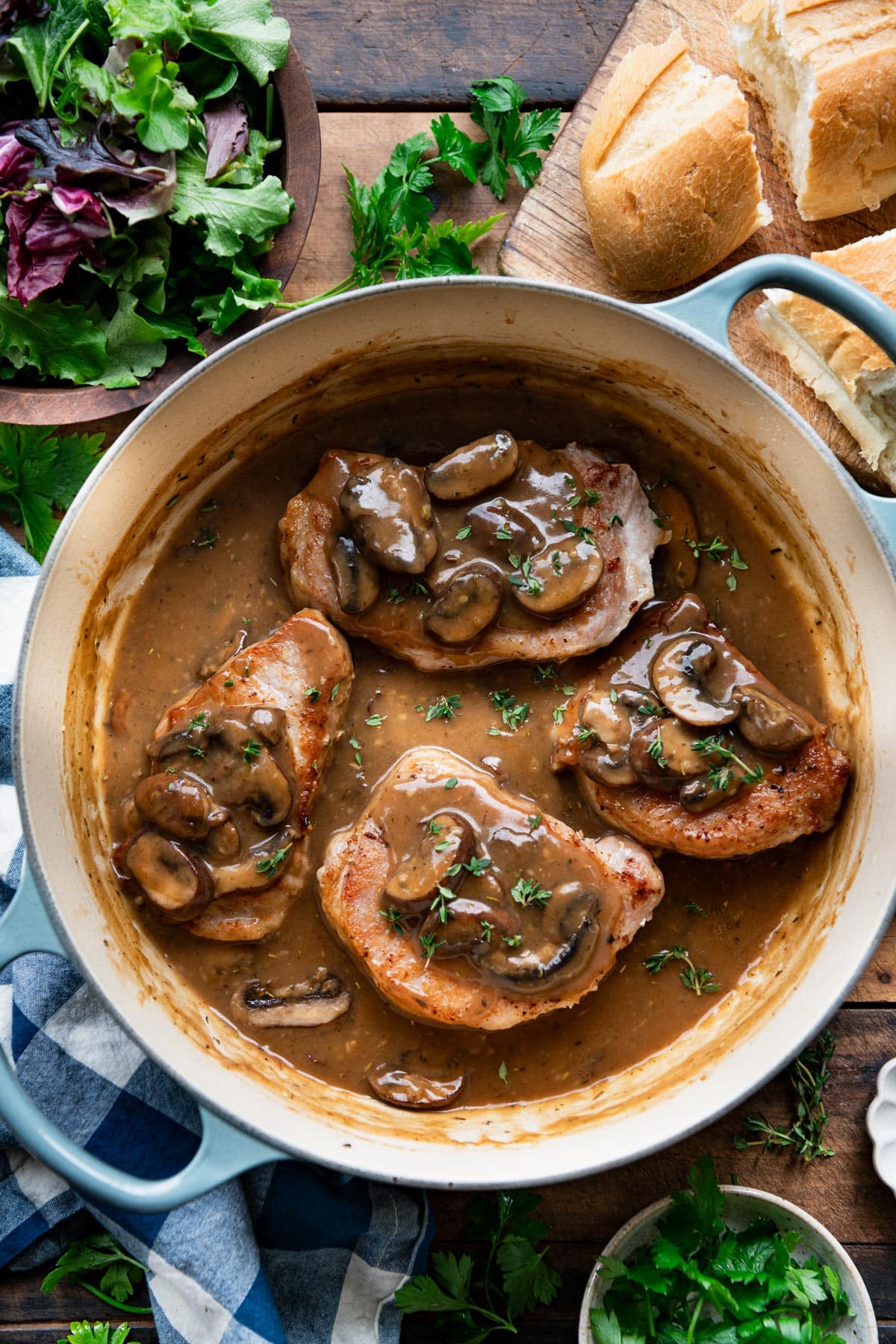 Overhead image of enamel cast iron dutch oven full of pork chops and mushroom gravy on a dinner table with salad and bread.