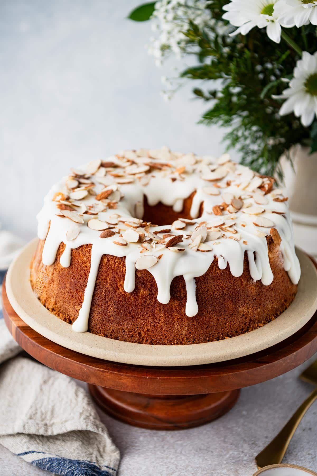 Side shot of a glazed almond pound cake on a white table with flowers in the background.