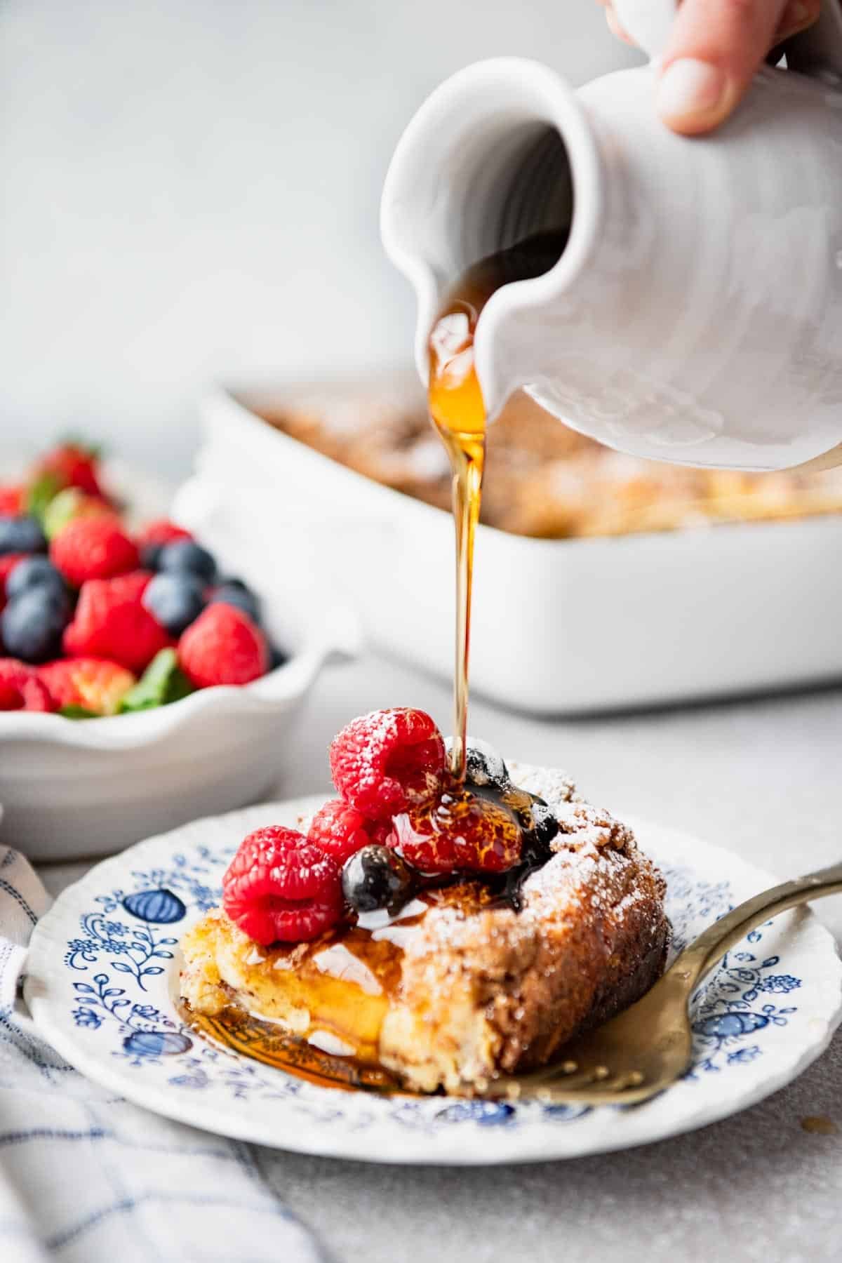 White pitcher drizzling syrup over a slice of overnight brioche french toast casserole with fresh berries.