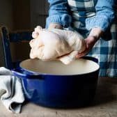 Hands holding a whole chicken to place it in a blue Dutch oven.