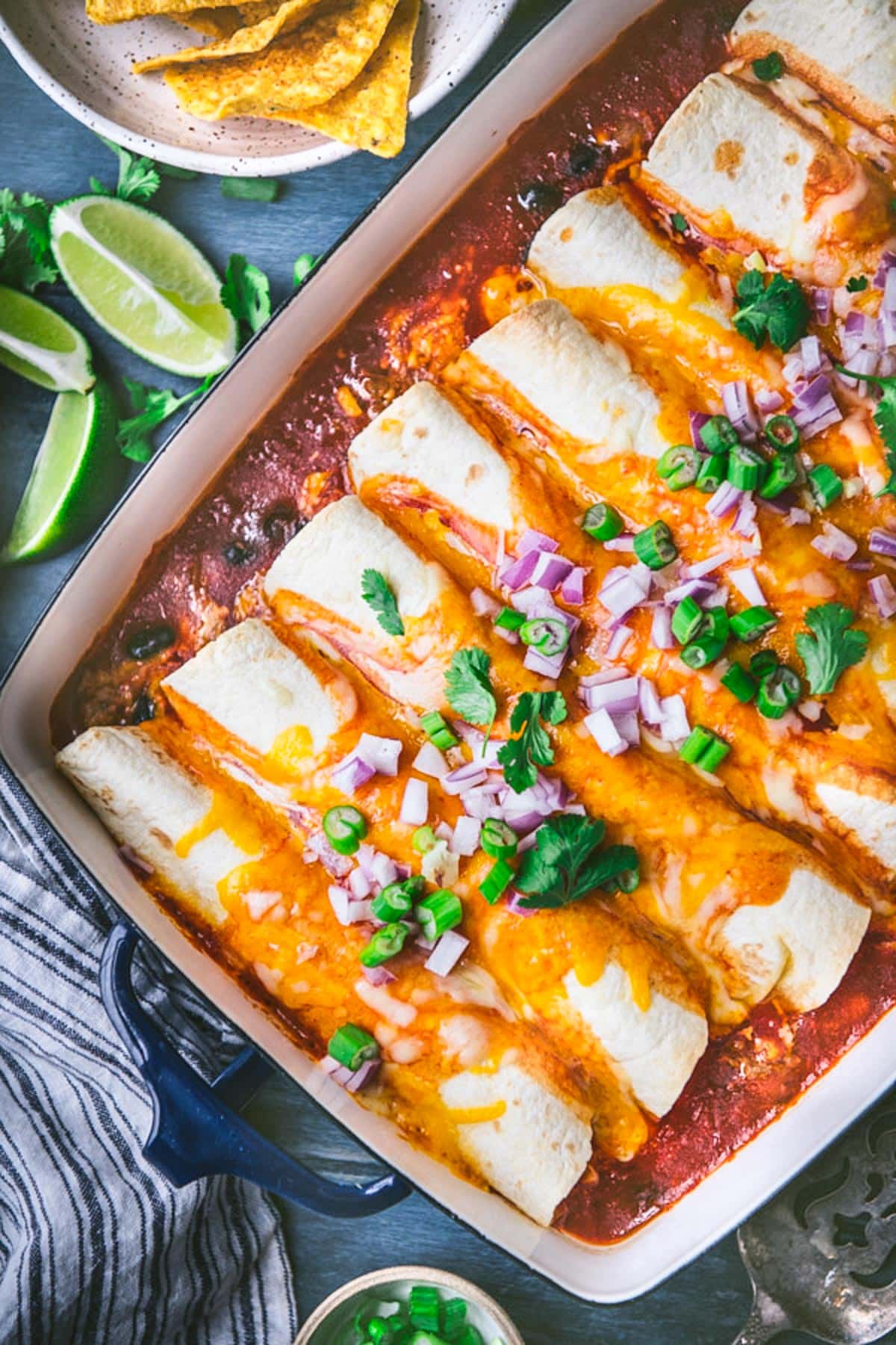 Close overhead shot of ground beef enchiladas in a baking dish with limes and tortilla chips and salsa on the side.