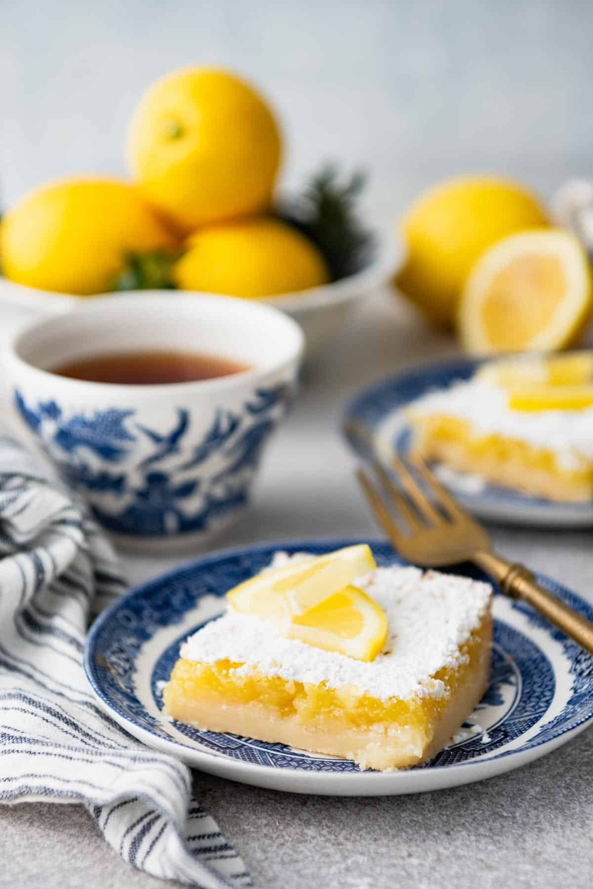 Side shot of lemon squares on plate with cups of tea on a white surface.
