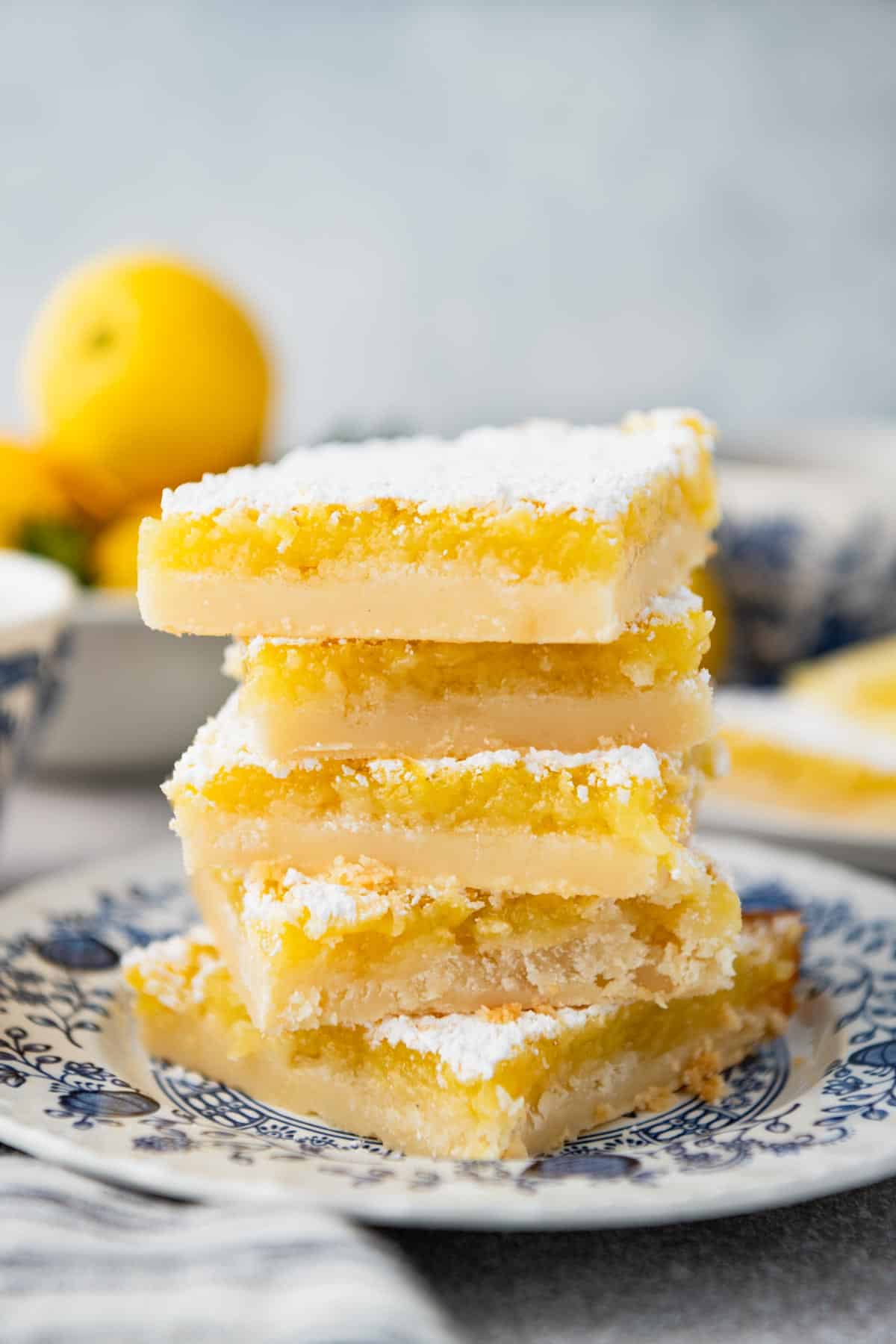 Close up side shot of a stack of lemon squares on a white table with fresh lemons in a bowl in the background.