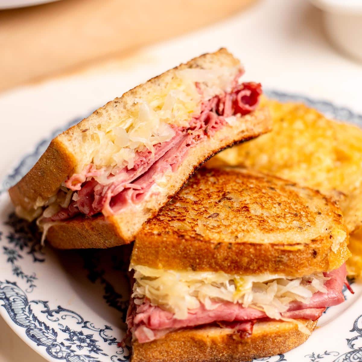 Square close up shot of a pastrami reuben sandwich on a blue and white plate.