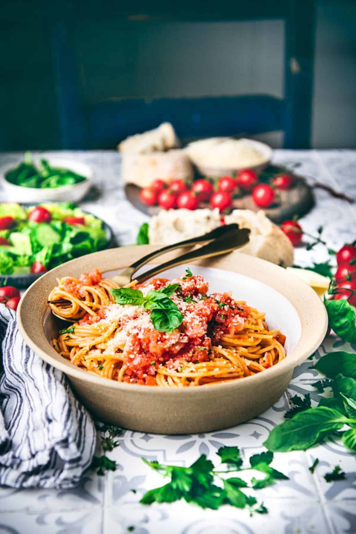Side shot of a bowl of pasta with pomodoro sauce on a dinner table with salad and bread in the background.