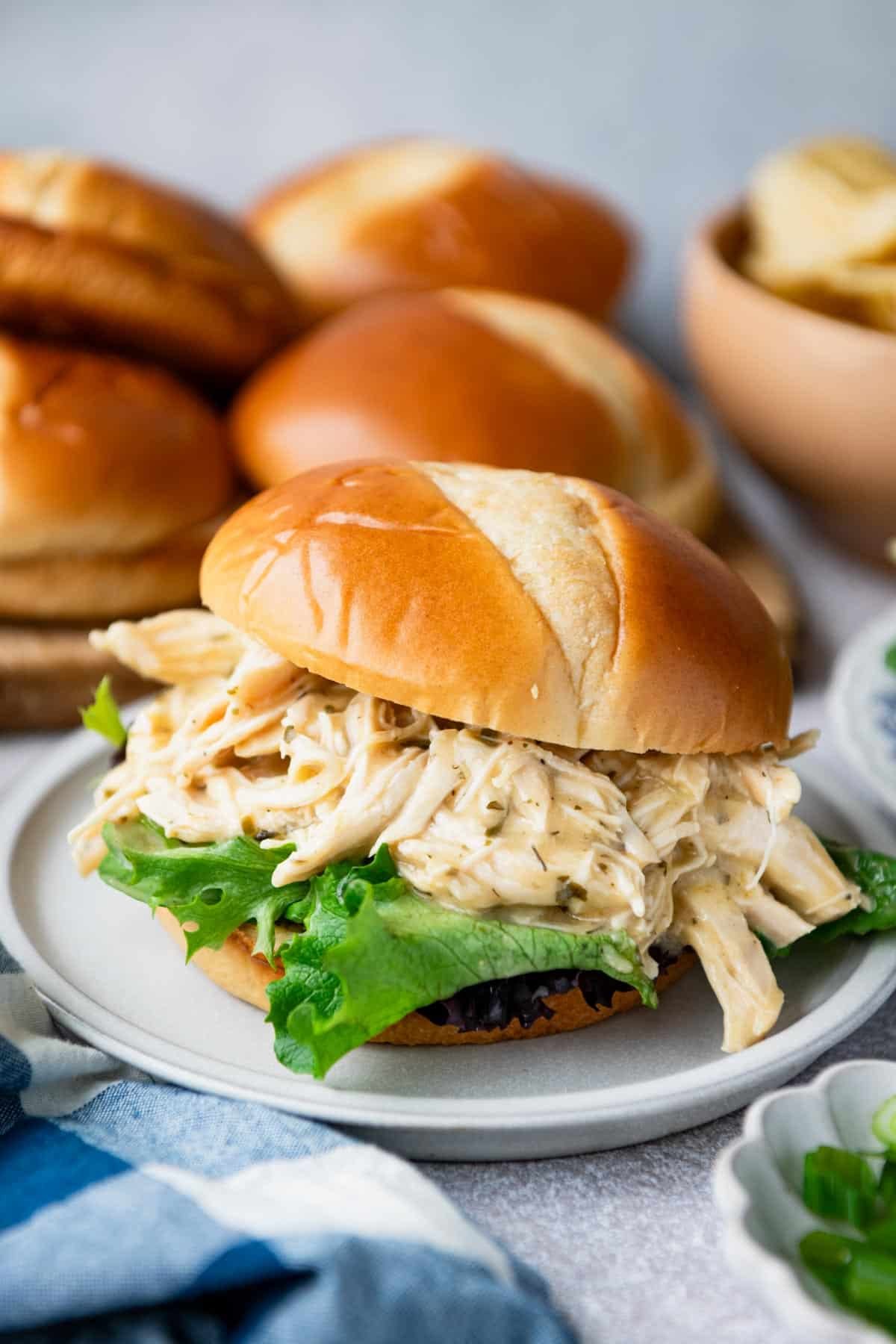 Ranch chicken crock pot sandwich on a white plate with rolls and chips in the background.