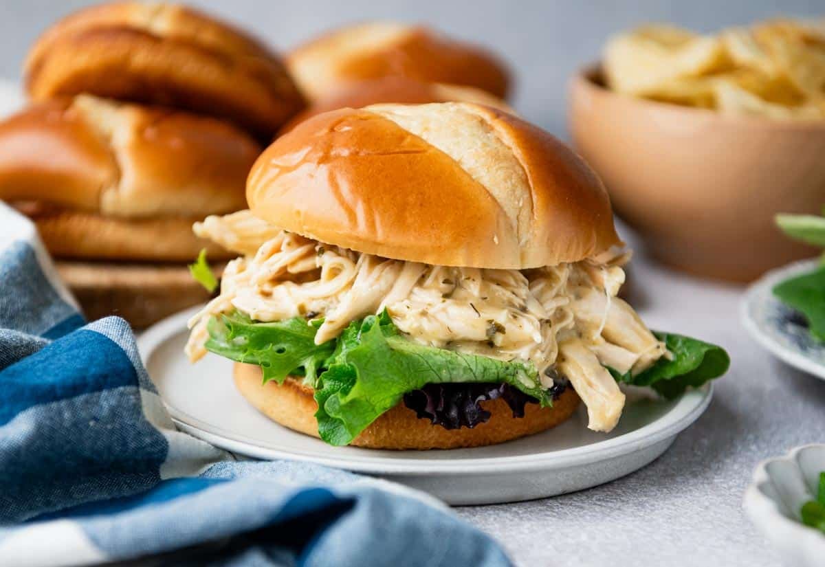Horizontal side shot of ranch chicken crock pot recipe served on a sandwich on a white plate with lettuce, buns, and chips in the background.