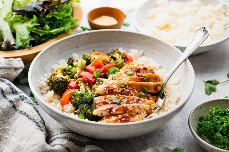 Horizontal side shot of baked teriyaki chicken in a bowl with rice and vegetables.