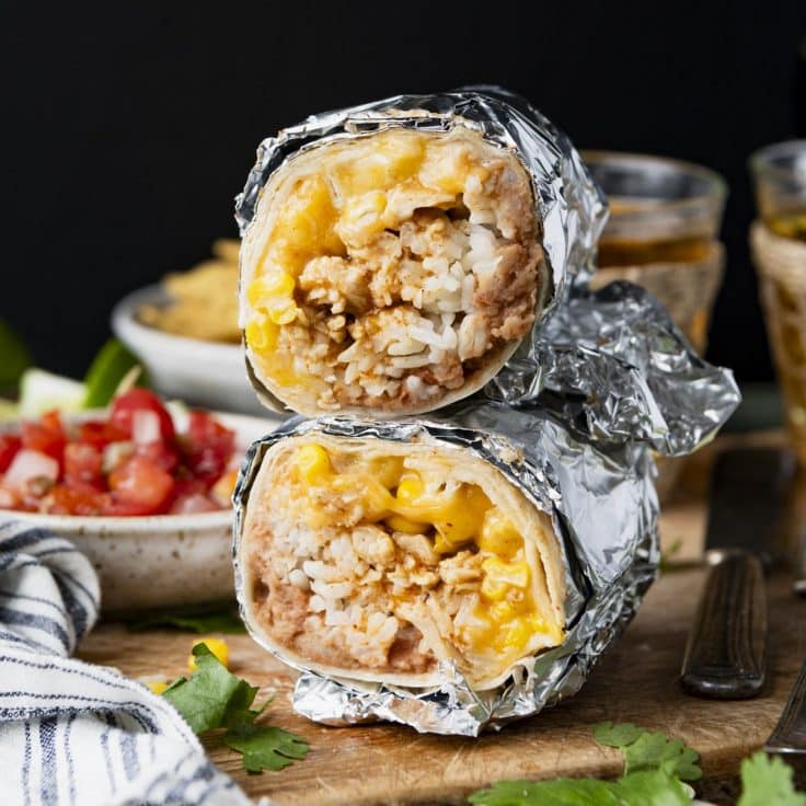 Stacked chicken burritos on a wooden board with chips and salsa in the background.