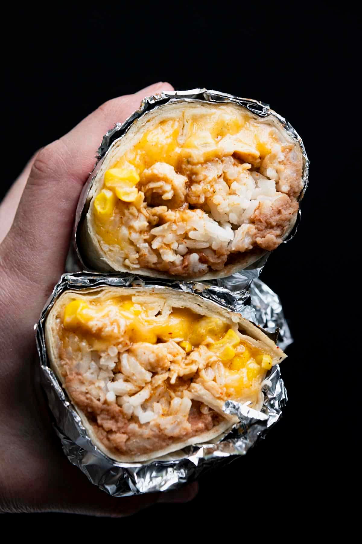 Hands holding a chicken burrito recipe wrapped in foil in front of a black background.