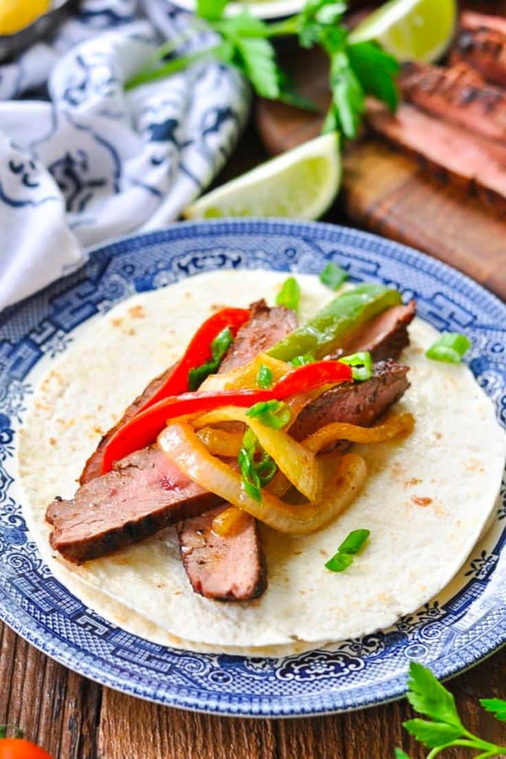 Flank steak fajitas on a blue and white plate with flour tortillas.