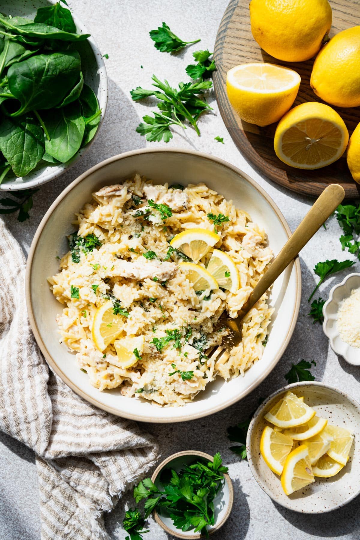 Bowl of lemon chicken orzo on a white table with spinach and a tray of lemons nearby.