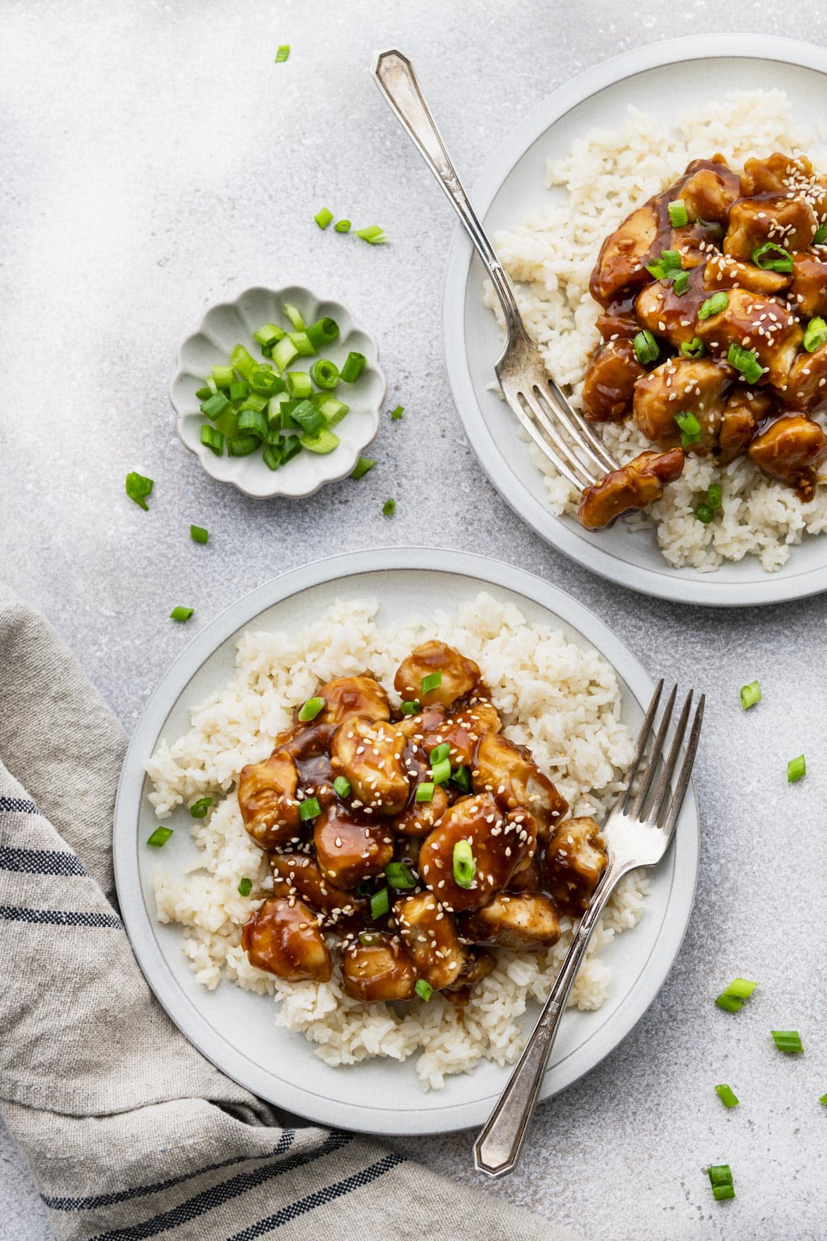 Overhead shot of two plates of sticky chicken on rice with green onions and sesame seeds on top.