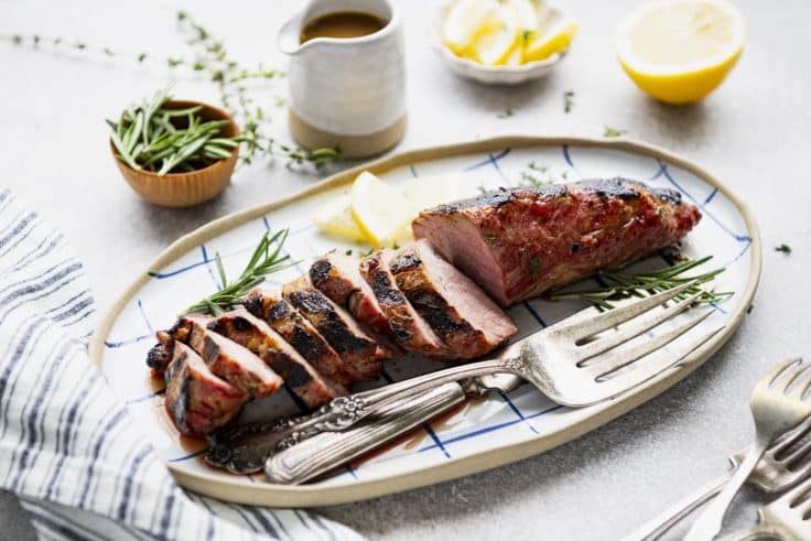 Horizontal image of the best pork tenderloin recipe served on a white table with a side of the best pork tenderloin marinade.